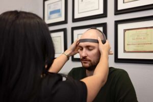 A woman places a headband-like device on a man’s head in an office setting, suggesting a Johnstown Chiropractor consultation; framed certificates hang on the wall in the background.
