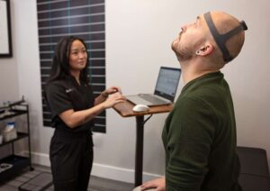A man wearing a headband device looks upward while standing in front of a laptop; a Johnstown Chiropractor and a woman in medical scrubs observe him in a clinical setting.