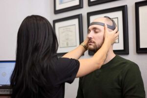 A Johnstown Chiropractor assists as a woman places a gray headband device on a man's head, who sits with his eyes closed. Several framed certificates hang on the wall behind them.