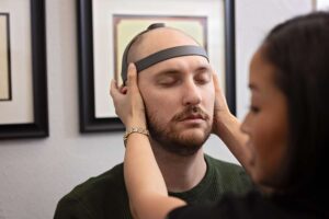 A woman places a headband-like device on a man's head as he sits with his eyes closed, possibly for a neurological assessment at a Johnstown Chiropractor's office. Framed certificates are displayed on the wall behind them.