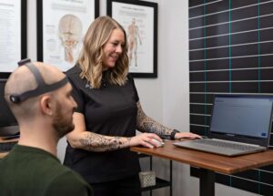 A woman in black scrubs smiles while operating a laptop on a standing desk. A man, possibly a Johnstown Chiropractor’s patient, sits nearby wearing a sensor headband. Medical charts are visible on the wall behind them.