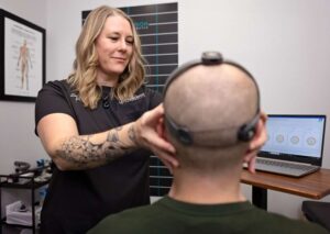 A Johnstown Chiropractor adjusts sensors on a person's bald head while monitoring data on a laptop in a clinical setting. Charts and anatomical diagrams are visible on the walls.