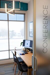 A modern conference room with a wooden table, two chairs, a desktop computer, and large windows—an inviting space where a Johnstown Chiropractor might consult. The glass door is partially open and etched vertically with CONFERENCE.