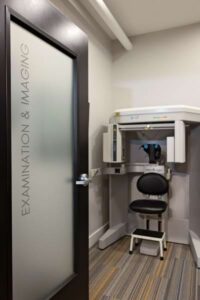 A small medical room with a frosted glass door labeled Examination & Imaging, often used by a Johnstown Chiropractor, features an imaging machine, black chair, step stool, white walls, and a striped floor.