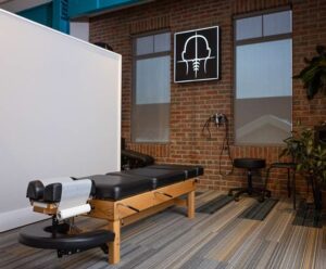 A Johnstown Chiropractor office featuring a black padded adjustment table, rolling stool, exam equipment on brick walls, large windows, plants, and a lit spine diagram sign for a welcoming and professional atmosphere.