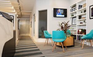 Modern office waiting area with teal chairs around a round wooden table, bookshelves with decor and books, a TV mounted on the wall, and a striped carpeted hallway leading to glass doors at this welcoming Johnstown Chiropractor.