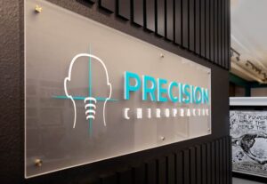 A wall sign reads “PRECISION CHIROPRACTIC” next to a simple outline of a head and spine on a transparent panel, mounted against a dark, textured wall—representing your trusted Johnstown Chiropractor.