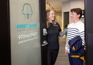 A smiling woman in a black uniform welcomes another smiling woman, who is holding a jacket, through the door of Precision Chiropractic clinic, a trusted Johnstown Chiropractor. The clinic’s name and contact info are visible on the glass door.