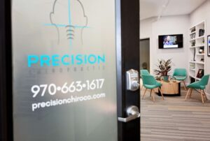 Glass door with Precision Chiropractic logo and contact info; inside, the Johnstown Chiropractor’s waiting area features teal chairs, a round wooden table, plants, bookshelves, and a wall-mounted TV.