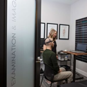 A person sits in a chair wearing a cap with sensors while another person stands nearby in a medical examination room. The door labeled Examination & Imaging is partially open.