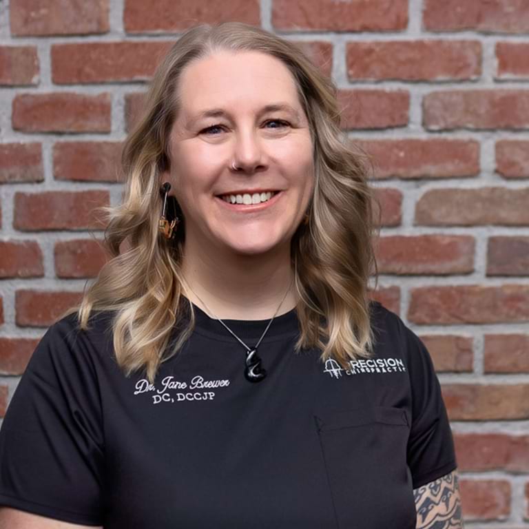 A woman with long, wavy blonde hair is smiling while standing in front of a brick wall. She is wearing a black shirt with embroidered text, a crescent moon necklace, and looks ready for her day at Precision Chiropractic.