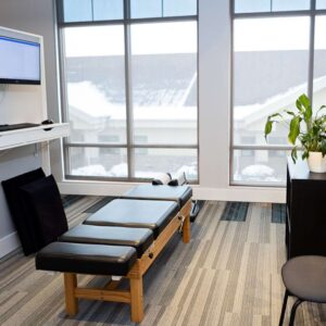 A modern Precision Chiro office with a black adjustment table, a potted plant on a cabinet, a chair, and large windows showing a snowy outdoor scene. A computer monitor is mounted on the wall.
