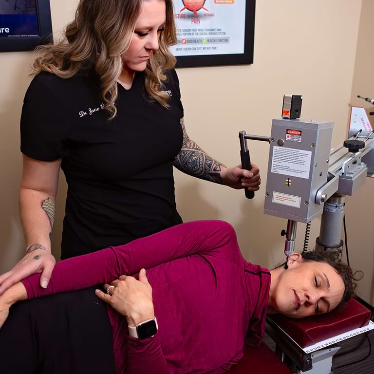 A Precision Chiropractic specialist adjusts a woman's lower back with a specialized tool while she lies on her side, eyes closed and relaxed. The chiropractor, wearing a black shirt and showing tattooed arms, ensures expert care.